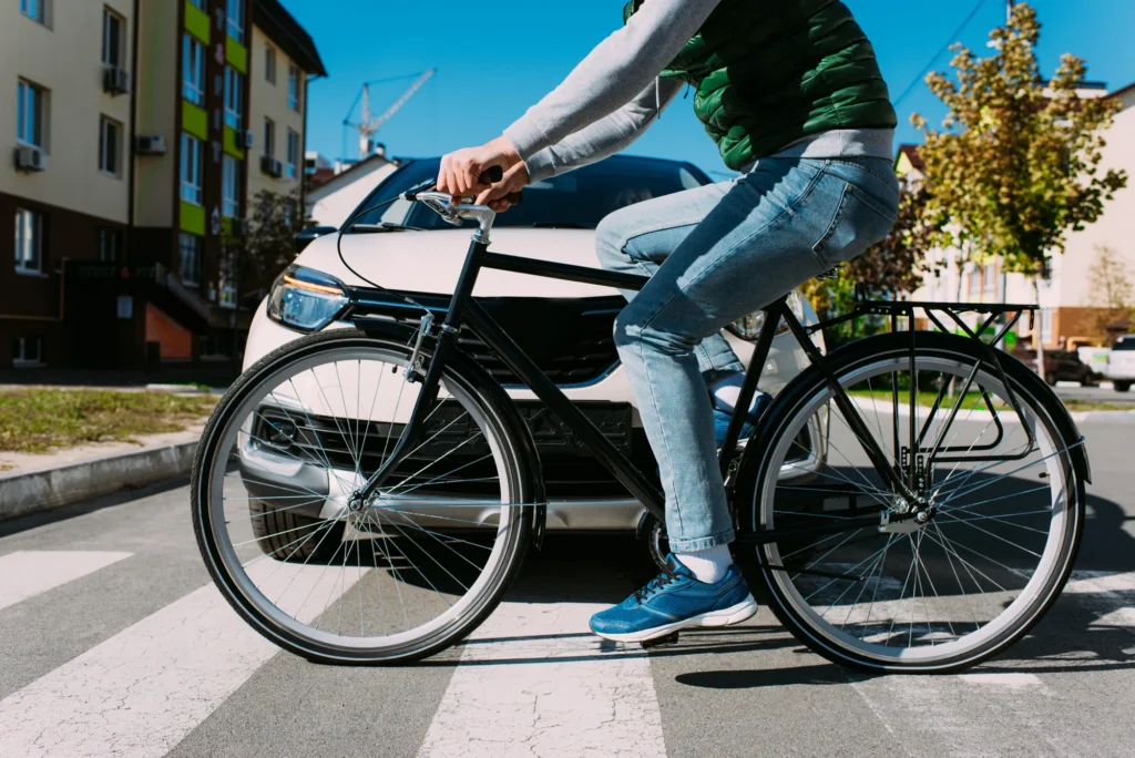 Urban street scene with a cyclist wearing a green vest and blue jeans riding a black bicycle in front of a white car under a blue sky; Martinez & Schill LLP referenced
