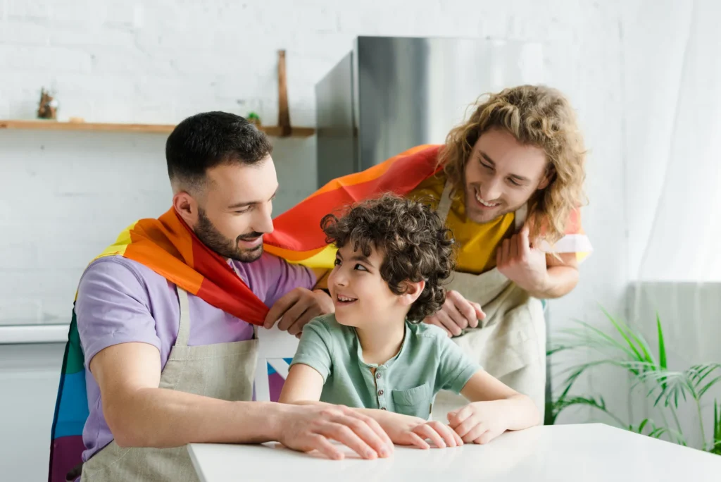 Colorful, smiling family moment in a bright kitchen: a cheerful boy sits at a table while two adults drape rainbow capes over him, capturing a playful, joyful bond.